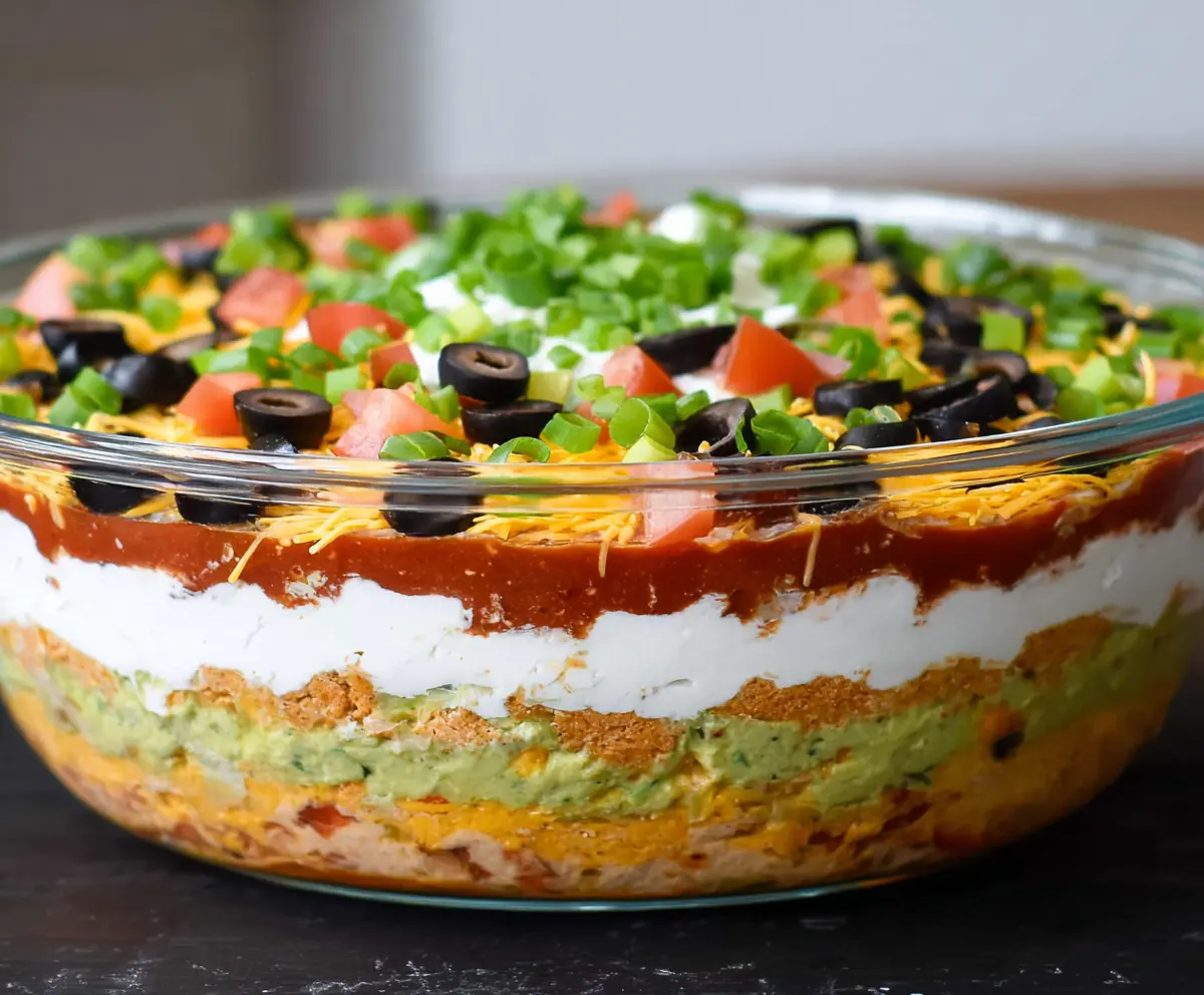 Delicious Ultimate 7 Layer Dip arranged in a clear bowl showcasing colorful ingredients like guacamole, sour cream, salsa, cheese, olives, and jalapenos.